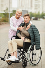 Fototapeta na wymiar Portrait of cheerful careful female social care worker with badge spending time with patient in wheelchair outdoors