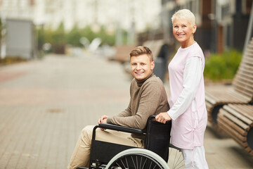 Positive mature blond nurse in uniform pushing wheelchair with smiling young patient in medical...