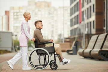 Content attractive qualified nurse in uniform strolling with disabled patient in city, she pushing...
