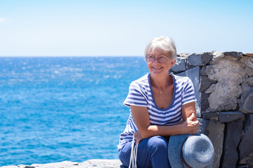 Smiling attractive senior woman dressed in blue sitting close to the sea in summer vacation enjoying sunny day