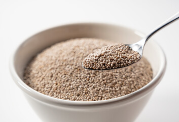 White chia seeds in a white bowl set against a white background. Scooping with a stainless steel spoon.