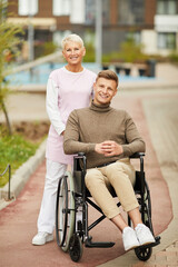 Fototapeta na wymiar Portrait of cheerful qualified nurse in medical uniform holding handles of wheelchair with handicapped man in yard of apartment building