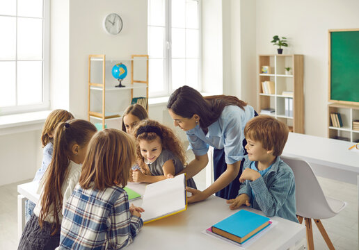 Professional Enthusiastic Young Female Teacher Reading Story With Diverse Group Of Happy Active Elementary School Pupils In Classroom. Learning Lesson, Back To School And Primary Education Concept