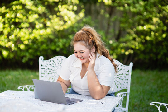 Young adult smiling fat Asian woman sitting at yard using laptop for buying and shopping online