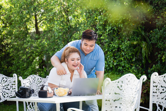 Young adult fat Asian happy couple sitting at yard using laptop for buy and online shopping