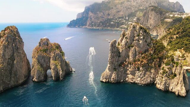 Aerial drone view of the Tyrrhenian sea coast of Capri, Italy. Rocky cliffs, blue water, floating boats, greenery, town in the distance
