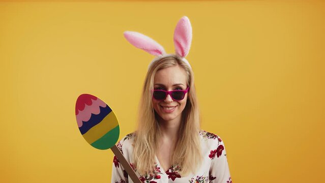Cool pretty blonde European girl dressed as easter bunny, holding cardboard colourful Easter egg. Yellow background studio shot. High quality 4k footage