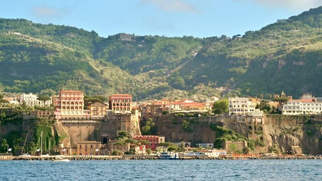 View of the Tyrrhenian sea coast in Sorrento, Italy. Rows of buildings, beach, cliffs, moving yacht, greenery. View from a boat