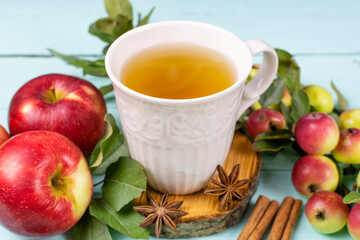 A white mug with tea on a wooden saw, ripe red apples, cinnamon sticks and anise on a blue (mint) background close-up. Composition.