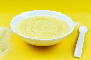 Corn porridge for a baby from ground cereals in a white bowl on a bright yellow background. Side view. Baby's first complementary food, baby nutrition.