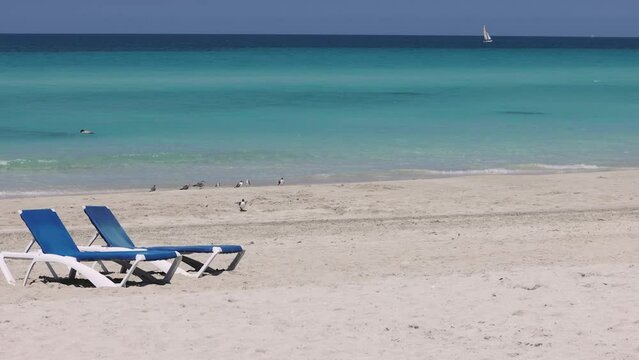 The beautiful beach front of the Cuban beach at Varadero in Cuba showing the waves and clean ocean waters with two deck chairs on the sandy beach with a sail boat filmed in 8k video quality