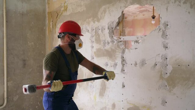 Contractor wrecks wall with sledgehammer making hole for rearrangement. Man doing manual dismantling and demolition works with big hammer hits for apartment renovation. Construction worker in uniform.
