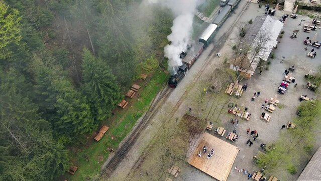 Aerial drone view of the steam train Mocanita in a valley on a railway station, resting tourists, lush forest and river around, Romania