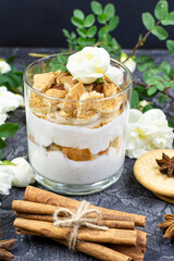 Cottage cheese-yogurt multi-layer dessert with crunchy cracker (cookies) and cocoa (cinnamon), white bush roses, anise and cinnamon sticks on a black background. Recipe, ingredients. Vertical photo.