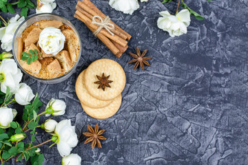 Fototapeta na wymiar Romantic breakfast. Multi-layer cottage cheese dessert with yogurt, cookies and cocoa in a glass glass, cinnamon sticks, anise, bushy white roses on a black (dark) background. Space for the text.