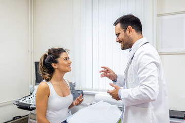 Fototapeta premium Male doctor consulting female patient at consultation. Professional physician wearing white coat talking to woman at appointment visit in clinic.