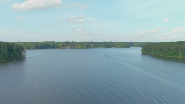 aerial footage of the rippling blue waters of Lake Acworth surrounded by lush green trees, grass and plants with a motor boat on the water with blue sky and clouds at Proctor Landing Park in Acworth