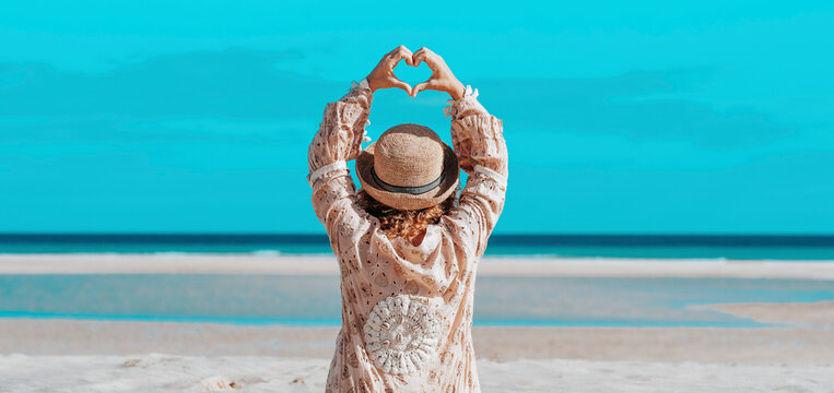 Travel and scenic beach destination concept lifestyle with woman with boho dress doing heart gesture with hands against a blue ocean and sky in background. Outdoo rleisure on vacation holiday