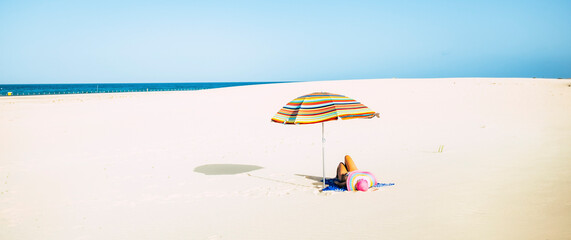 Woman tourist laying on the sand at the beach with straw hat and colorful umbrella enjoying the sun in summer holiday vacation leisure activity. People relaxing under the sun at empty tropical beach