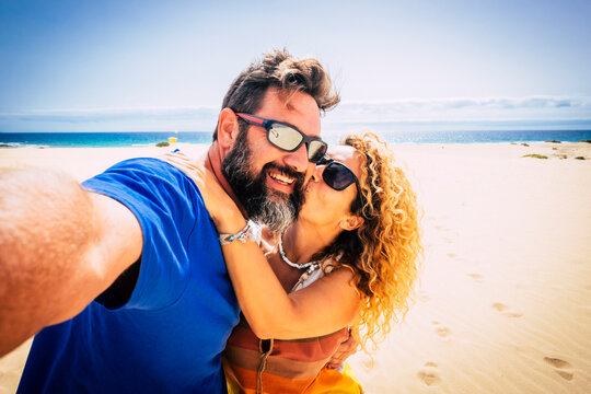 Adult couple of tourist take selfie picture at the beach with sand and blue ocean and sky in background. Hapy man and woman enjoying summer holiday vacation together. Travel lifestyle people