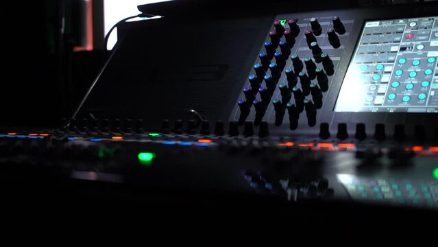A panning shot of a soundboard. The buttons and faders of the soundboard are bright and are contrasted against the dark lighting.