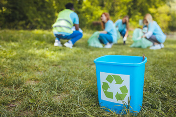 Recycling bin in focus. In the field. With a sign on it. In the background, volunteers are cleaning...