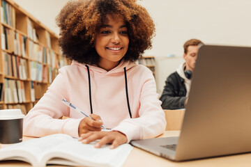 Smiling girl writing in a book while sitting at desk with laptop. Female student studying at...