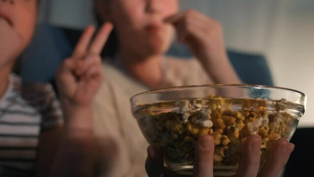 Plate is submissive in hands. Sister girls are watching TV. eating popcorn sitting on couch. Children take popcorn from plate in front of TV eat. Children watch their favorite TV, eat salted popcorn