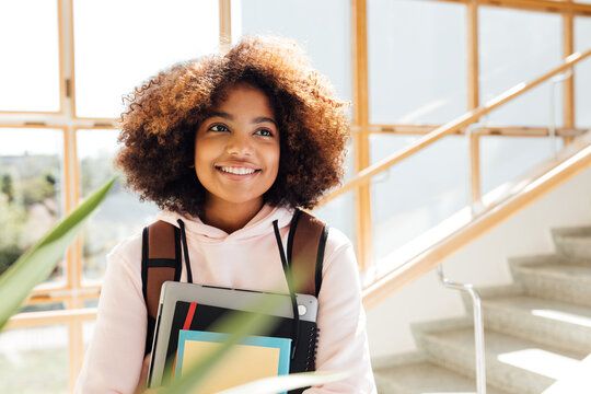 Cheerful girl with backpack holding textbooks. Young female smiling and looking away in school.