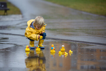 Beautiful funny blonde toddler boy with rubber ducks and colorful umbrella, jumping in puddles and...