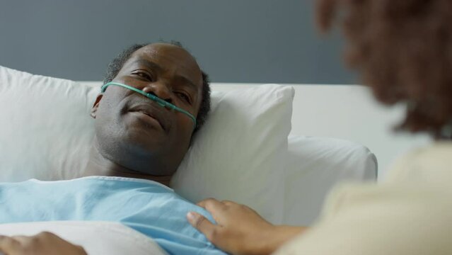 Tilt up over the shoulder shot of sick African American man with nasal cannula lying on hospital bed and talking with loving wife as she holding his hands and stroking shoulder