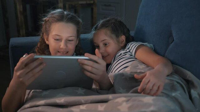 Two cute girls lying on couch under warm blanket in room are looking at screen of digital tablet.Modern digital entertainment technologies for children. Children learn through digital tablet, have fun