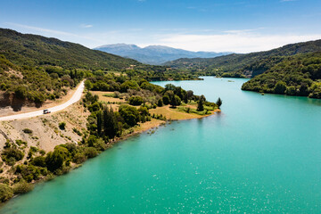 Obraz na płótnie Canvas Roadtrip: Aerial view on a blue lake with road winding along the shore and an 4x4 vehicle parking close to it