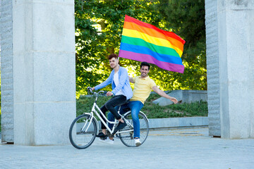 Young gay men couple riding a bike holding a gay pride rainbow flag outdoors