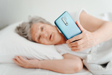 Obraz na płótnie Canvas Surprised senior woman holding mobile phone, emotionally looking at smartphone screen, shocked mature woman lying in bed in bedroom. Selective focus on phone