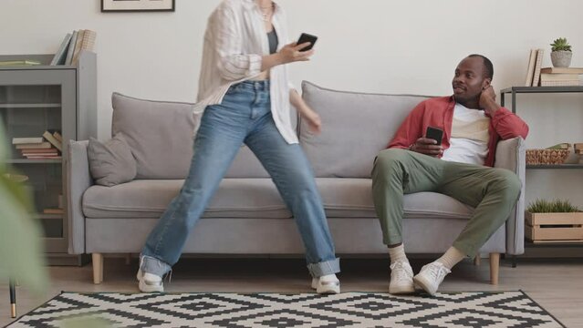 Stab shot of young cheerful multiethnic couple laughing at funny memes and videos while scrolling feed on social media, sitting together on sofa in living room