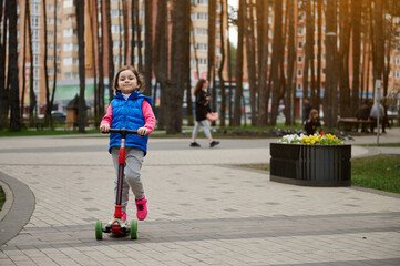 Full length portrait of an European cute child girl riding a kick scooter in the park on a sunny day in early autumn or spring. happy childhood concept