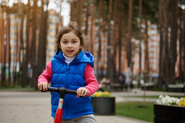 Adorable child girl riding a kick scooter in the city park on a sunny day in early autumn or spring. Healthy and active lifestyle, leisure activity