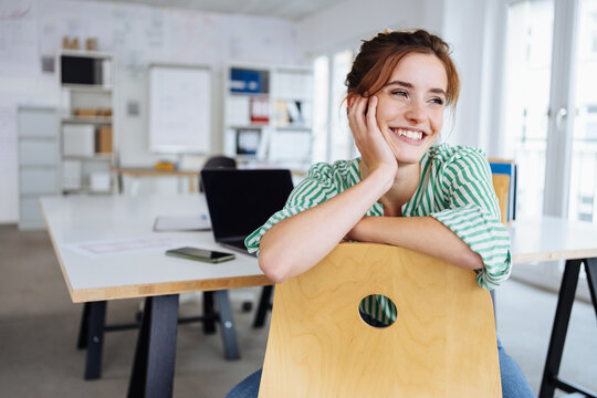 Happy young business woman leans on her desk chair and looks to the side laughing