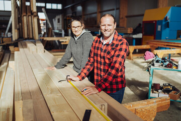 Two carpenters stand next to a stack of wood and measure the length