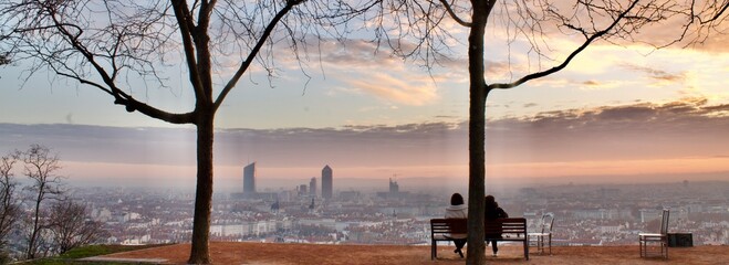 Panoramic picture with 2 people sitting on a bench admiring a wonderful sunrise over the city of...