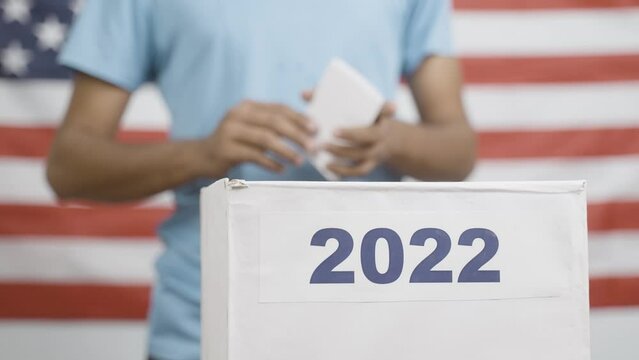 Man placing ballot paper into 2022 ballot box with us or american flag as background - concept of 2022 election, voting, responsibility and democracy.
