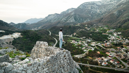 A young guy stands on the edge of the ruined Haj Nehai fortress against the backdrop of a valley and mountains. The guy poses against the backdrop of the mountains of Montenegro. Fortress in Sutomore