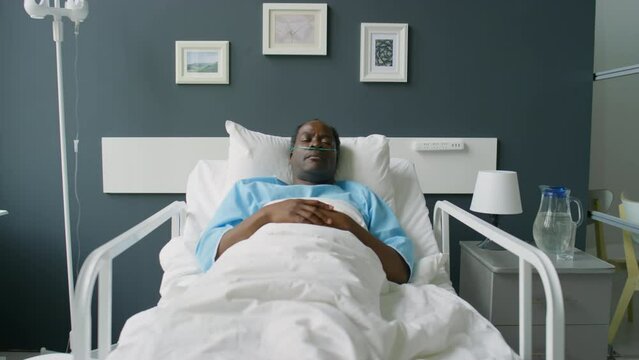 Zoom in shot of unconscious African American man with nasal cannula lying on bed with eyes closed in hospital ward