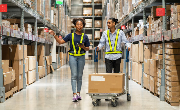 African American woman walks with a tablet and an Asian man picks up boxes in a cart to deliver to customers walking in a warehouse at a furniture wholesaler.