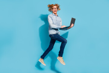 Fototapeta na wymiar Full length photo of excited pretty young guy dressed jeans shirt eyewear jumping high chtting modern gadget isolated blue color background