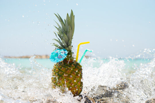 Fresh pineapple fruit cocktail with a straw and a blue umbrella stands on a rock in a spray of sea water on a tropical background. Summer vacation and summer vacation concept. 