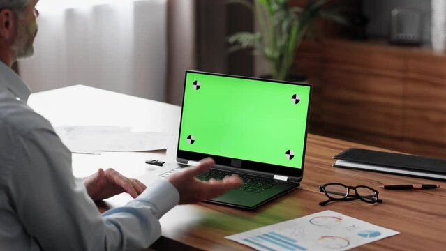 Businessman has video call on laptop green screen with tracking markers. Male worker talking and listening during video conference with chroma key computer screen. Over shoulder laptop screen view.