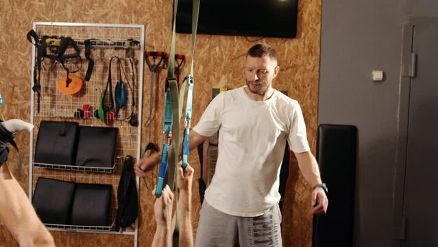 Trainer monitors exercise. Men training arms with trx fitness straps in gym doing push ups in air train upper body chest shoulders pecs triceps. People exercising in gym, physical education concept