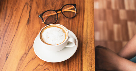 Cup of cappuccino coffee with latte and glasses of a woman on a wooden bar counter in a sunlit cafe in the morning.